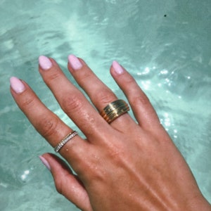 Pu&ograve; includere: Una mano con smalto rosa chiaro sulle unghie indossa due anelli d'oro. L'anello sull'indice &egrave; una fascia spessa con pi&ugrave; scanalature. L'anello sul medio &egrave; una fascia sottile con piccoli ciottoli trasparenti.