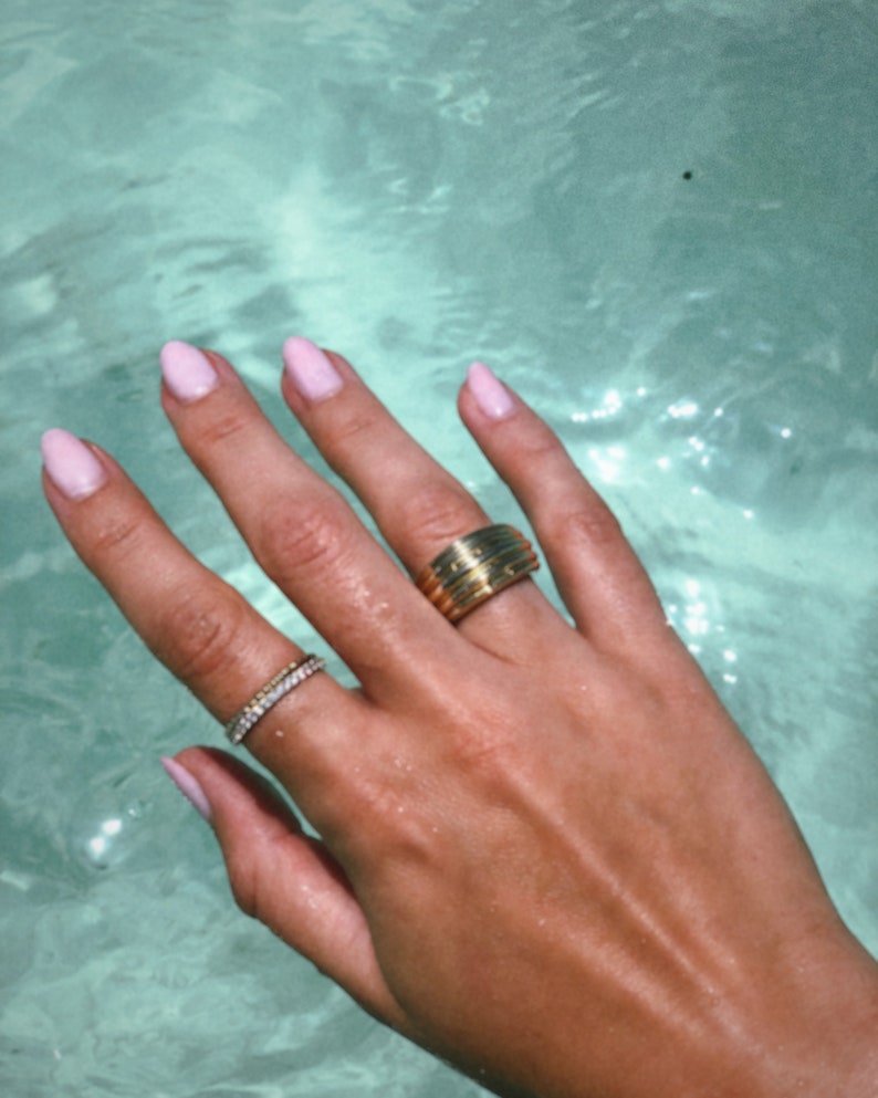 Pu&ograve; includere: Una mano con smalto rosa chiaro sulle unghie indossa due anelli d'oro. L'anello sull'indice &egrave; una fascia spessa con pi&ugrave; scanalature. L'anello sul medio &egrave; una fascia sottile con piccoli ciottoli trasparenti.