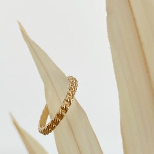 Può includere: Un anello d'oro con un design intrecciato. L'anello è su uno sfondo marrone chiaro.