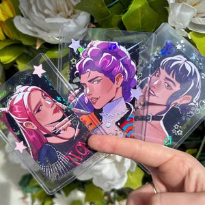 Op de afbeelding: Drie doorzichtige plastic kaarthouders met kleurrijke illustraties van gestileerde personages. Elke kaart toont een ander personage met unieke kapsels en kleding. De kaarten zijn versierd met sterren en andere kleine vormen.