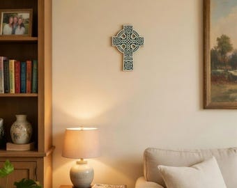 May include: A Celtic cross wall hanging with a green and white design is mounted on a cream-colored wall. The cross is the focal point of the image, with a lamp and side table in the foreground and a bookshelf to the left.