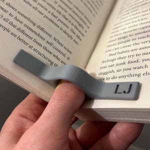 May include: A grey 3D printed bookmark with the initials "LJ" printed on it. The bookmark is designed to hold a page open in a book.