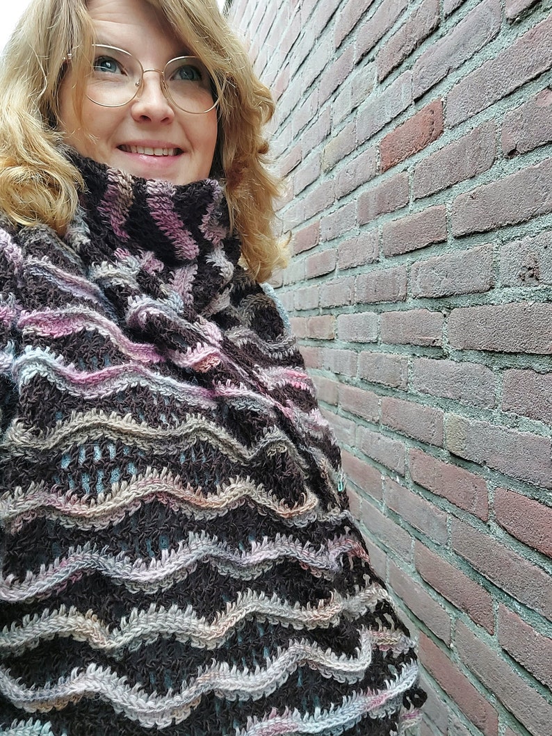 Pu&ograve; includere: Una donna indossa uno scialle all'uncinetto marrone, rosa e blu con un motivo ondulato.