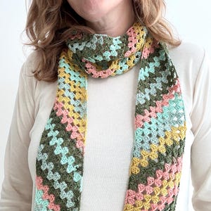 May include: A crochet scarf featuring a diagonal granny stitch pattern in a multi-colored palette of olive green, soft yellow, pale pink, and light mint green. The scarf is constructed with an open, airy texture and hangs in two long vertical panels.