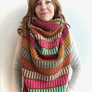 May include: A handmade crochet shawl in a vibrant palette of brown, pink, teal, and white. The shawl has a striped pattern with a textured, ribbed design. The triangular shape is draped around the neck and shoulders.