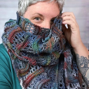 Pu&ograve; includere: Una persona indossa uno scialle all'uncinetto multicolore con un motivo traforato. Lo scialle &egrave; realizzato con una variet&agrave; di tonalit&agrave; di blu, verde, marrone e bianco.