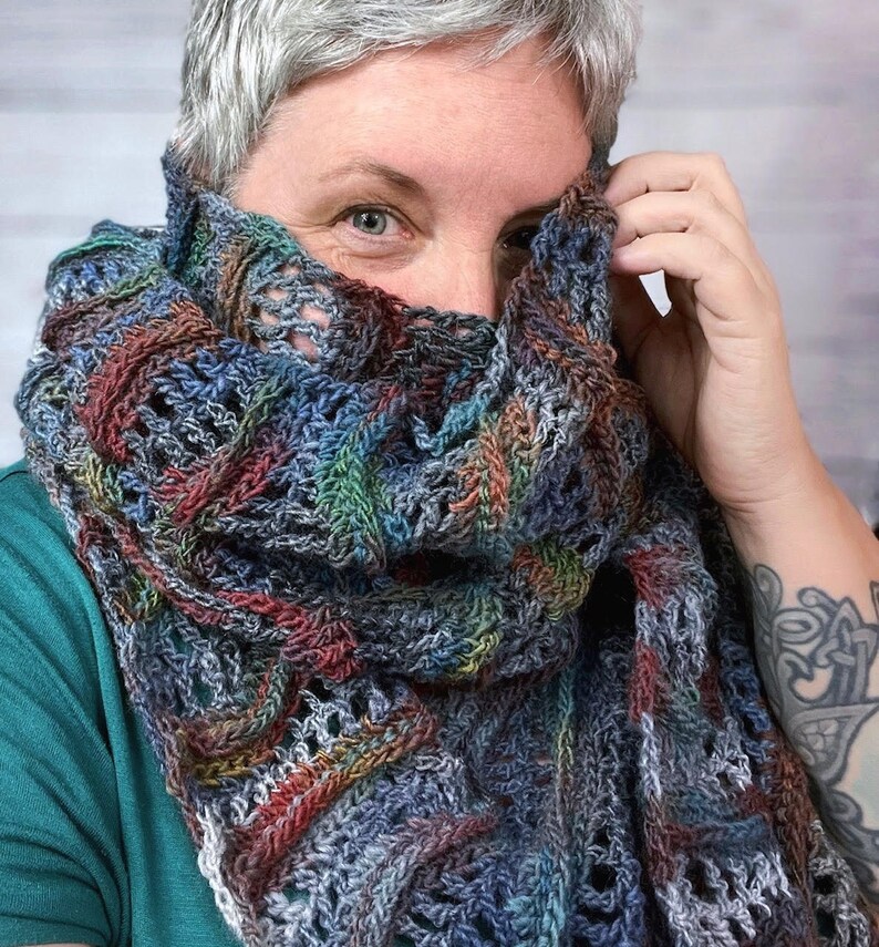 Pu&ograve; includere: Una persona indossa uno scialle all'uncinetto multicolore con un motivo traforato. Lo scialle &egrave; realizzato con una variet&agrave; di tonalit&agrave; di blu, verde, marrone e bianco.