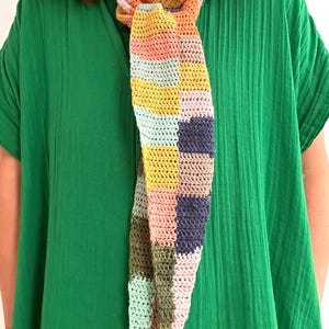 May include: A colorful crocheted scarf with a patchwork design in shades of pink, green, yellow, and blue. The scarf is tied around the neck of a person wearing a green shirt.