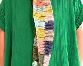 May include: A colorful crocheted scarf with a patchwork design in shades of pink, green, yellow, and blue. The scarf is tied around the neck of a person wearing a green shirt.
