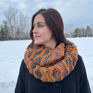 Pu&ograve; includere: Una donna indossa un cappotto nero con una sciarpa infinita marrone, blu e arancione. La sciarpa &egrave; realizzata a uncinetto con un motivo ondulato.