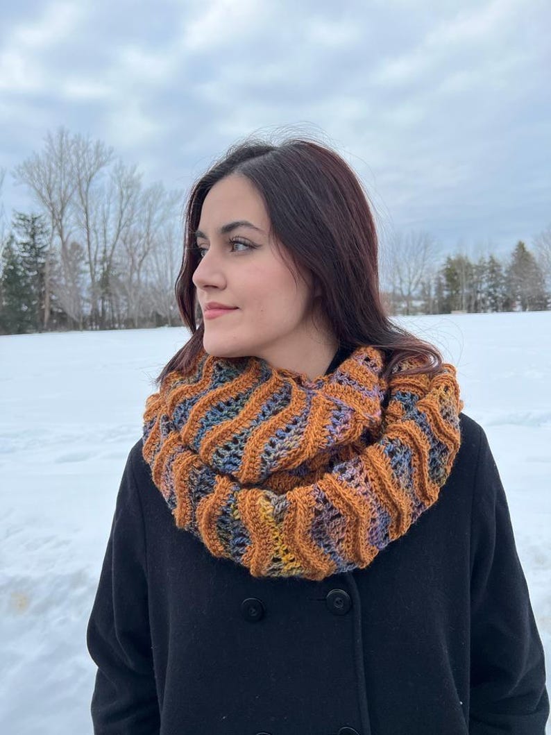 Pu&ograve; includere: Una donna indossa un cappotto nero con una sciarpa infinita marrone, blu e arancione. La sciarpa &egrave; realizzata a uncinetto con un motivo ondulato.