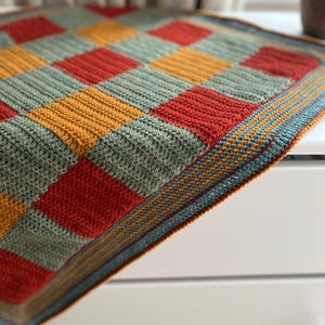 May include: A crocheted blanket with a checkered pattern in shades of green, red, and yellow. The blanket has a blue, yellow, and orange border.