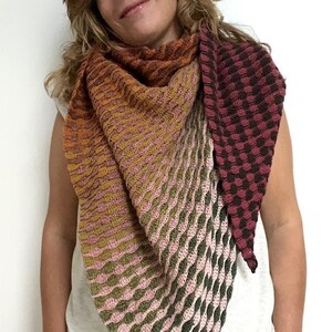 May include: A crocheted shawl with a gradient of colors, starting with orange and transitioning to green. The shawl has a textured pattern and is worn by a person.