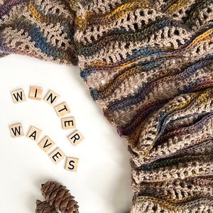 Pu&ograve; includere: Un primo piano di una sciarpa all'uncinetto in tonalit&agrave; marrone, beige e blu. La sciarpa &egrave; realizzata con un punto testurizzato e presenta un motivo ondulato. Le parole "WINTER WAVES" sono scritte in lettere nere su piastrelle bianche.