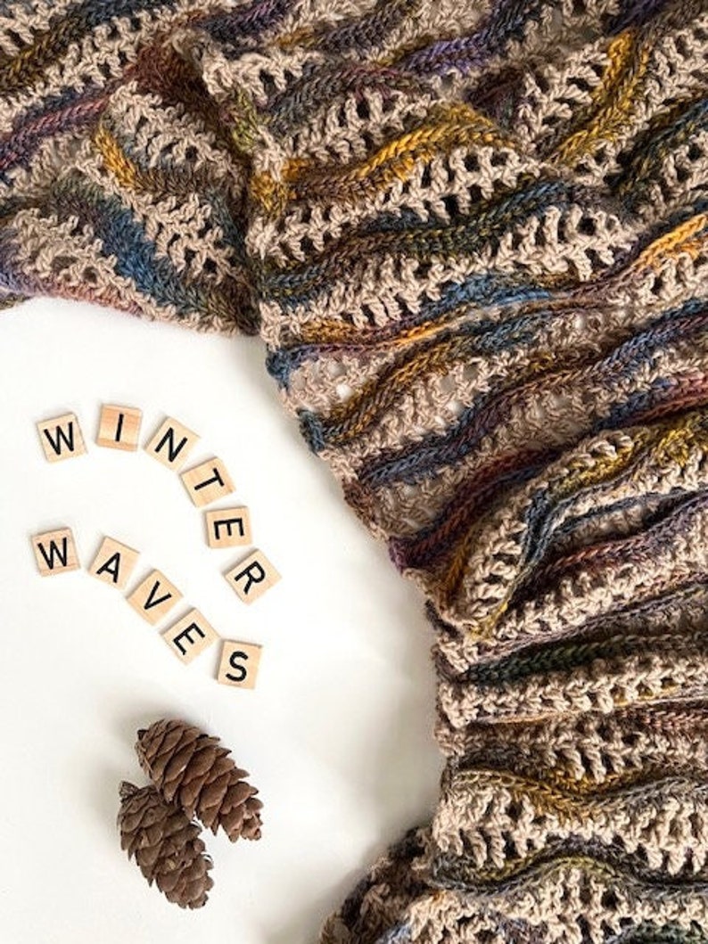 Pu&ograve; includere: Un primo piano di una sciarpa all'uncinetto in tonalit&agrave; marrone, beige e blu. La sciarpa &egrave; realizzata con un punto testurizzato e presenta un motivo ondulato. Le parole "WINTER WAVES" sono scritte in lettere nere su piastrelle bianche.