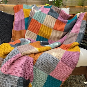 Op de afbeelding: Een kleurrijk gehaakt deken met vierkanten in tinten roze, oranje, geel, teal, grijs en bruin. Het deken is over een stoel gegooid.