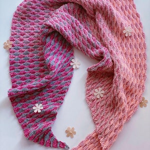 May include: A pink and gray crocheted shawl with a textured pattern. The shawl is draped over a white surface with small decorative flowers.