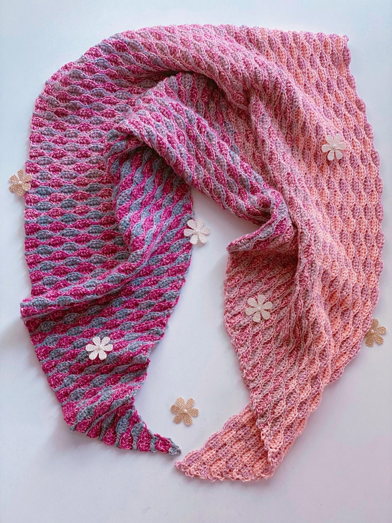 May include: A pink and gray crocheted shawl with a textured pattern. The shawl is draped over a white surface with small decorative flowers.