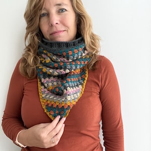 May include: A woman wearing a rust-coloured long-sleeved shirt with a colourful crocheted triangular scarf. The scarf has a grey, teal, pink, orange, and yellow colour scheme.