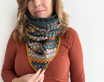 May include: A woman wearing a rust-colored long-sleeved shirt with a colorful crocheted triangular scarf. The scarf has a gray, teal, pink, orange, and yellow color scheme.