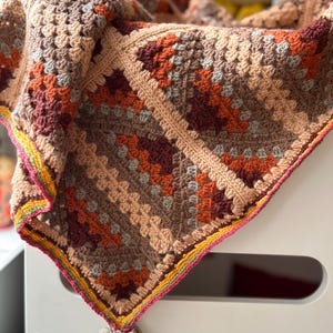 May include: A crocheted blanket with a colorful geometric pattern in shades of brown, orange, and pink. The blanket has a fringe border and a pom-pom on one corner.