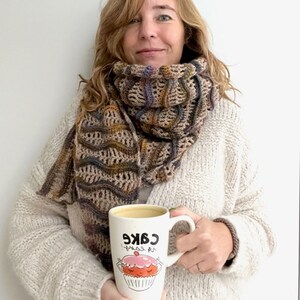 Pu&ograve; includere: Una donna indossa una sciarpa all'uncinetto marrone e beige con un maglione bianco e tiene in mano una tazza bianca con il testo "Cake is my therapy" e un'illustrazione di cupcake di cartone animato.