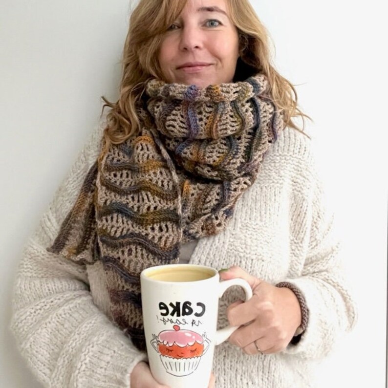 Pu&ograve; includere: Una donna indossa una sciarpa all'uncinetto marrone e beige con un maglione bianco e tiene in mano una tazza bianca con il testo "Cake is my therapy" e un'illustrazione di cupcake di cartone animato.