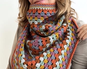 May include: A colourful crocheted scarf with a geometric pattern in shades of orange, green, blue, and brown. The scarf is folded in a triangle shape and has a crocheted border.