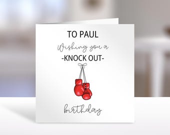 May include: A square white greeting card featuring the black text TO PAUL Wishing you a -KNOCK OUT- birthday. Below the text is an illustration of a pair of red boxing gloves tied together with a thin gray string and a small bow at the top.
