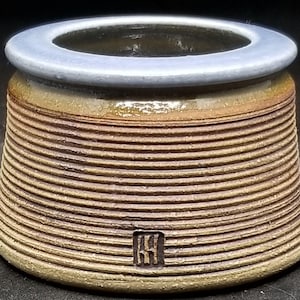 May include: A low, wide ceramic vessel with a tapered, ribbed exterior in shades of brown and tan. The top rim is smooth and light gray. A small, square maker's mark with vertical lines is embossed near the base of the textured, ridged body.
