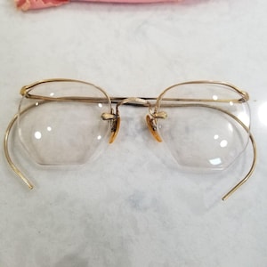 May include: Antique gold-coloured spectacles with clear, octagonal lenses. The frames have a delicate wire design and small, orange nose pads. The temples curve around the ears. The glasses are set against a white background.