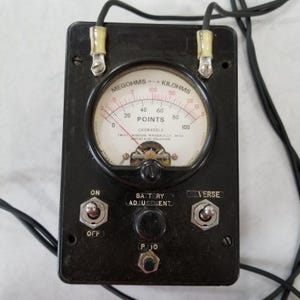 Puede incluir: Un probador eléctrico vintage negro con un dial redondo etiquetado como "Megohms - Kilohms" y "Points". El dial tiene una aguja que apunta a 60. El probador tiene tres interruptores de palanca etiquetados como "On", "Off", "Reverse" y una perilla etiquetada como "Ajuste de batería".