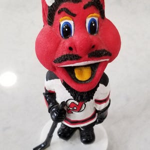 May include: A red devil mascot figurine wearing a white and red hockey jersey with the New Jersey Devils logo. The mascot has a black beard, mustache, and horns. The figurine is holding a black hockey stick.