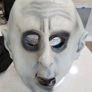 May include: A pale gray Halloween mask with dark eye sockets and a downturned mouth. The mask has a wrinkled forehead and prominent cheekbones. Text is visible through the eye holes.