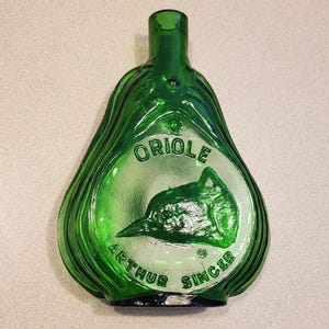 Può includere: Una bottiglia di vetro verde con corpo a forma di pera e collo rotondo. La bottiglia ha un'etichetta bianca con le parole "ORIOLE ARTHUR SINGER" stampate su di essa. La bottiglia presenta un'immagine in rilievo di un uccello.