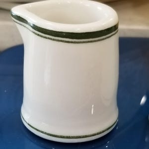 May include: A small, white ceramic creamer with a spout. The creamer has a dark green band around the top and bottom rims. The creamer is on a blue surface.