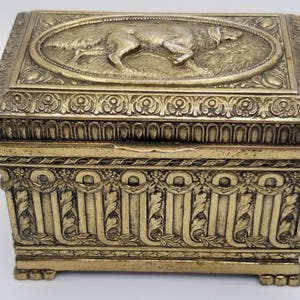 May include: A rectangular, antique brass box with ornate detailing. The lid features an oval relief of a dog in a field. The box has decorative floral and geometric patterns, with small feet. A vintage decorative storage item.