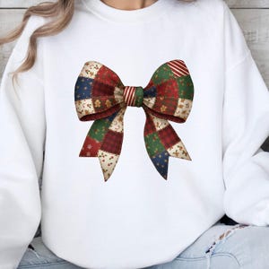 May include: A white sweatshirt featuring a large, colorful bow design. The bow is composed of patchwork squares in red, green, blue, and cream, with festive patterns like gingerbread men and snowflakes. The bow's center has red and white stripes.