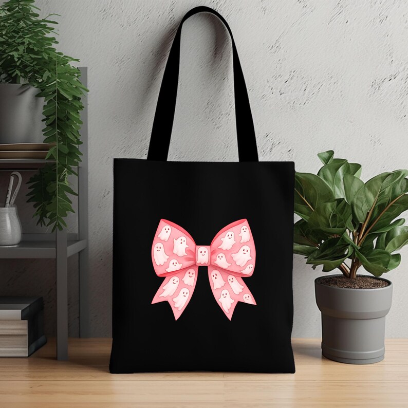 May include: Black tote bag featuring a pink bow design filled with white ghost illustrations. The tote bag has black handles and is made of a durable fabric. The bow design adds a playful touch to the bag.