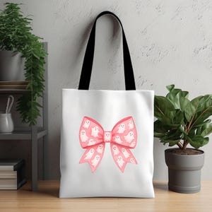 May include: White tote bag with a pink bow design filled with white ghost illustrations. The bag has black handles and is suitable for carrying various items. The design is whimsical and perfect for Halloween or a spooky-themed event.