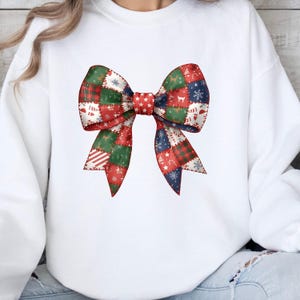 May include: White sweatshirt featuring a large, colorful bow graphic. The bow is composed of patchwork squares in red, green, and blue, with festive patterns like snowflakes and stars. The bow's center is a solid red with white stars.