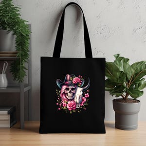 May include: Black tote bag with a pink and white skull wearing a cowboy hat and surrounded by pink flowers. The skull is half human and half cow skull.