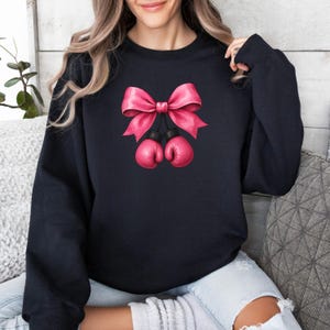 May include: Black sweatshirt featuring a pink bow and two pink boxing gloves. The bow is large and detailed, with a satin-like appearance. The boxing gloves are small and round, hanging below the bow.