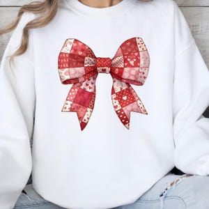 Pode incluir: Camisola branca com um grande laço decorativo. O laço é um design de patchwork em tons de vermelho e rosa, adornado com padrões de corações. O design do laço está centrado na frente da camisola, criando um tema de Dia dos Namorados.