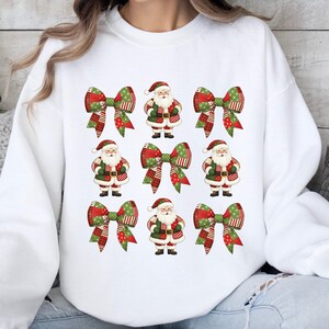 Op de afbeelding: Witte sweatshirt met een herhaald patroon van kerstmanfiguren en kerststrikken. De strikken hebben rood-groene ruit- en stippatronen. De kerstman draagt een rode hoed en een groen pak.