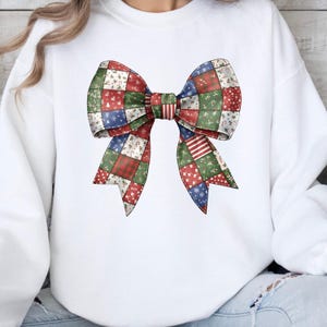 May include: White sweatshirt featuring a large, colorful bow design. The bow is made of patchwork squares in red, green, and blue, with festive patterns. A cozy and stylish top for the holiday season.
