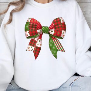 May include: White sweatshirt featuring a large, colorful bow design. The bow is a patchwork of red, green, and white patterns, including plaid, polka dots, and festive holiday motifs like gingerbread men and snowflakes. The bow is centered on the chest.