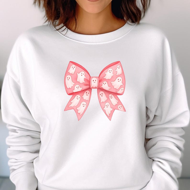 May include: White sweatshirt featuring a large pink bow with ghost illustrations. The bow is the focal point, with the ghosts adding a playful touch. The design is centered on the front of the sweatshirt, creating a festive and whimsical look.