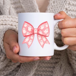 May include: White ceramic mug featuring a pink bow design filled with smiling ghost illustrations. The mug has a curved handle and is held by a person wearing a light-colored sweater. The design is perfect for Halloween or spooky season.