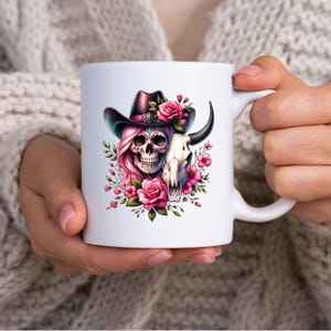 May include: White ceramic mug with a colorful illustration of a sugar skull wearing a cowboy hat and holding a bull skull. The skull is surrounded by pink flowers.